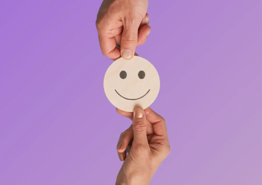 Deux mains tiennent un smiley en bois sur fond violet, symbole de feedback positif en entreprise